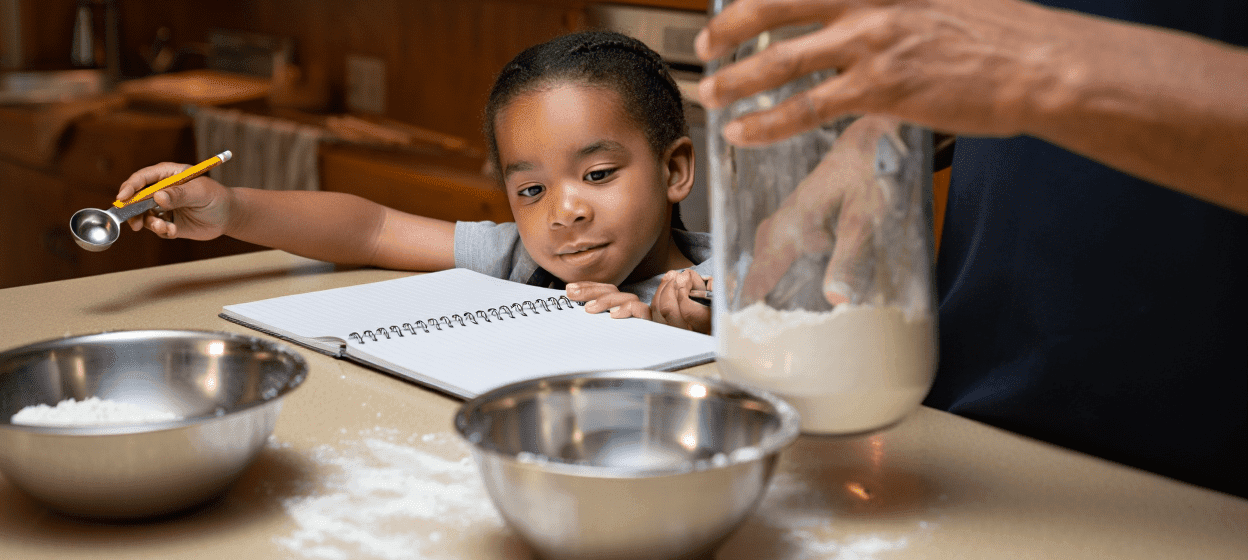 Child holding a measuring spoon, adult reaching into flour, with bowls and notebook.